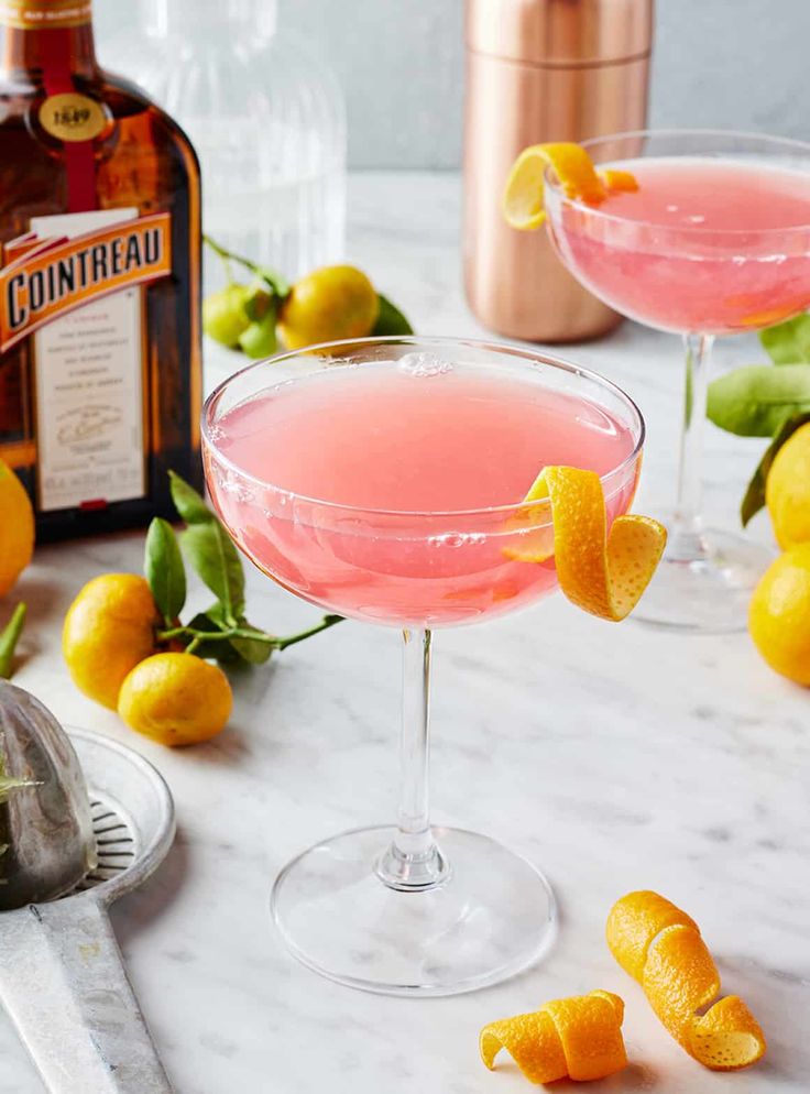 Pink cocktail in a glass with orange twist garnish on marble table, surrounded by fresh citrus and a Cointreau bottle.