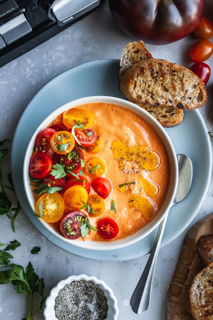 Colorful tomato soup topped with cherry tomatoes and herbs, served with crusty bread and black pepper.