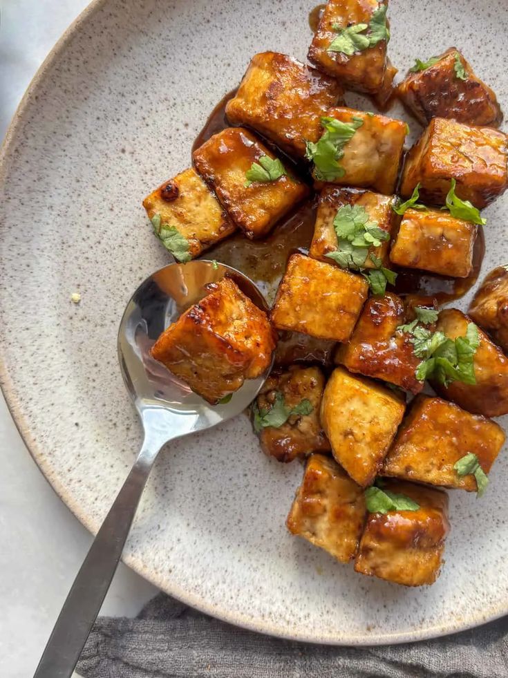 Crispy tofu cubes with glaze and herbs on a ceramic plate beside a spoon, perfect vegan dish.