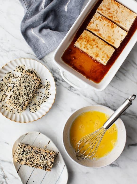 Tofu preparation with sesame coating, egg mixture, and marinade on a marble countertop.
