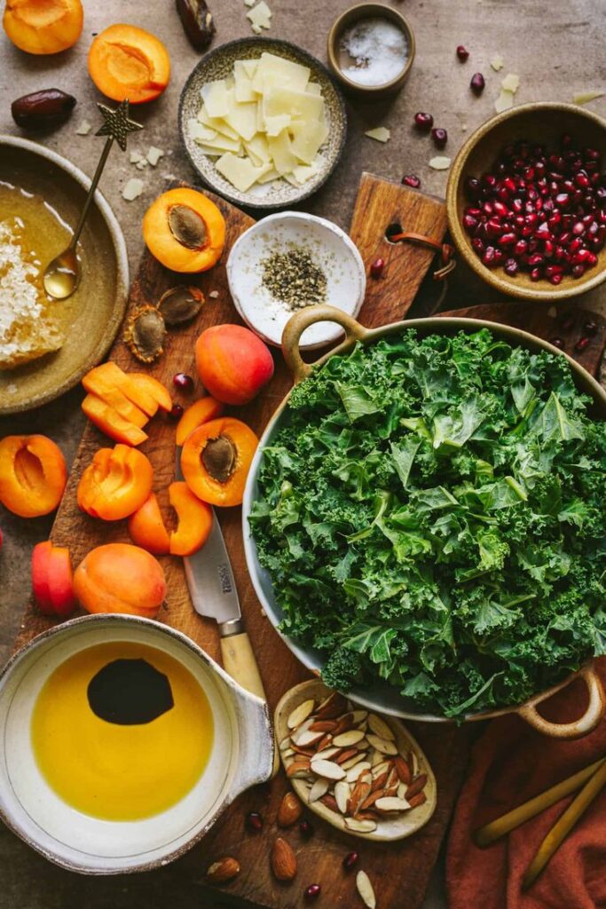 Fresh salad ingredients including kale, apricots, almonds, pomegranate, cheese, honey, and olive oil on a rustic table.
