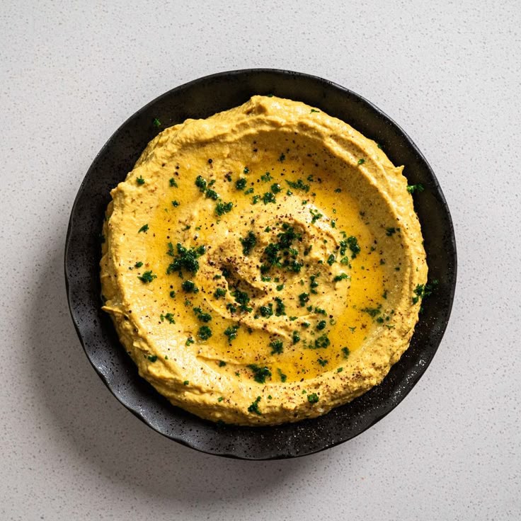 Creamy hummus topped with olive oil and herbs in a black bowl on a white background.