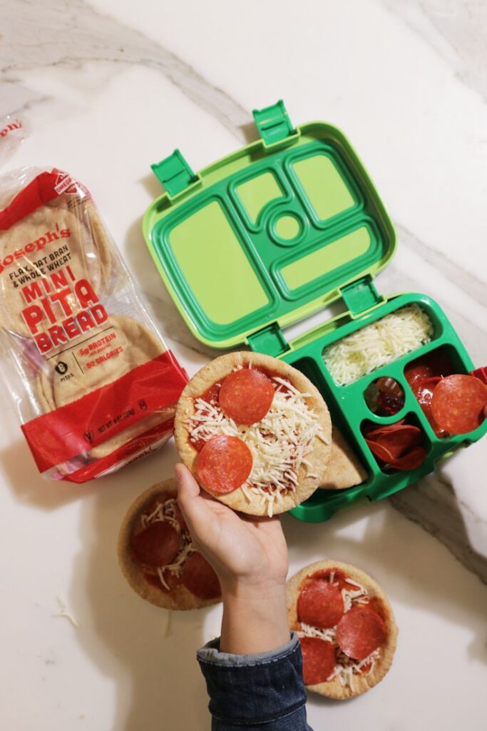 Mini pita bread pizzas with pepperoni and cheese beside a green lunchbox on a marble surface.