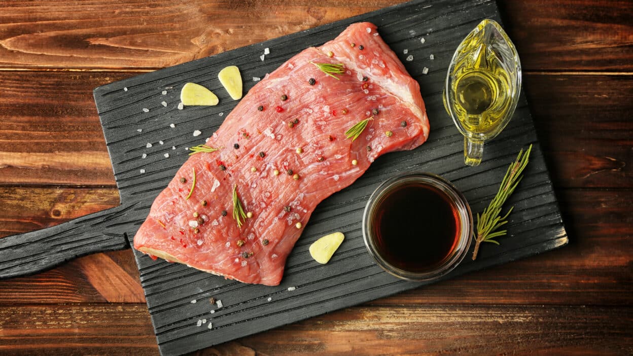 Raw beef steak seasoned with herbs, salt, and pepper on a black cutting board with olive oil and rosemary sprigs.