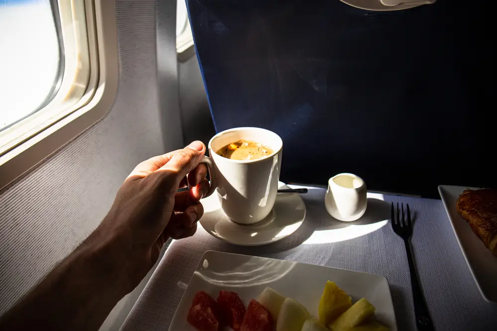 A person enjoying coffee and breakfast fruit on an airplane, with sunlight streaming through the window.