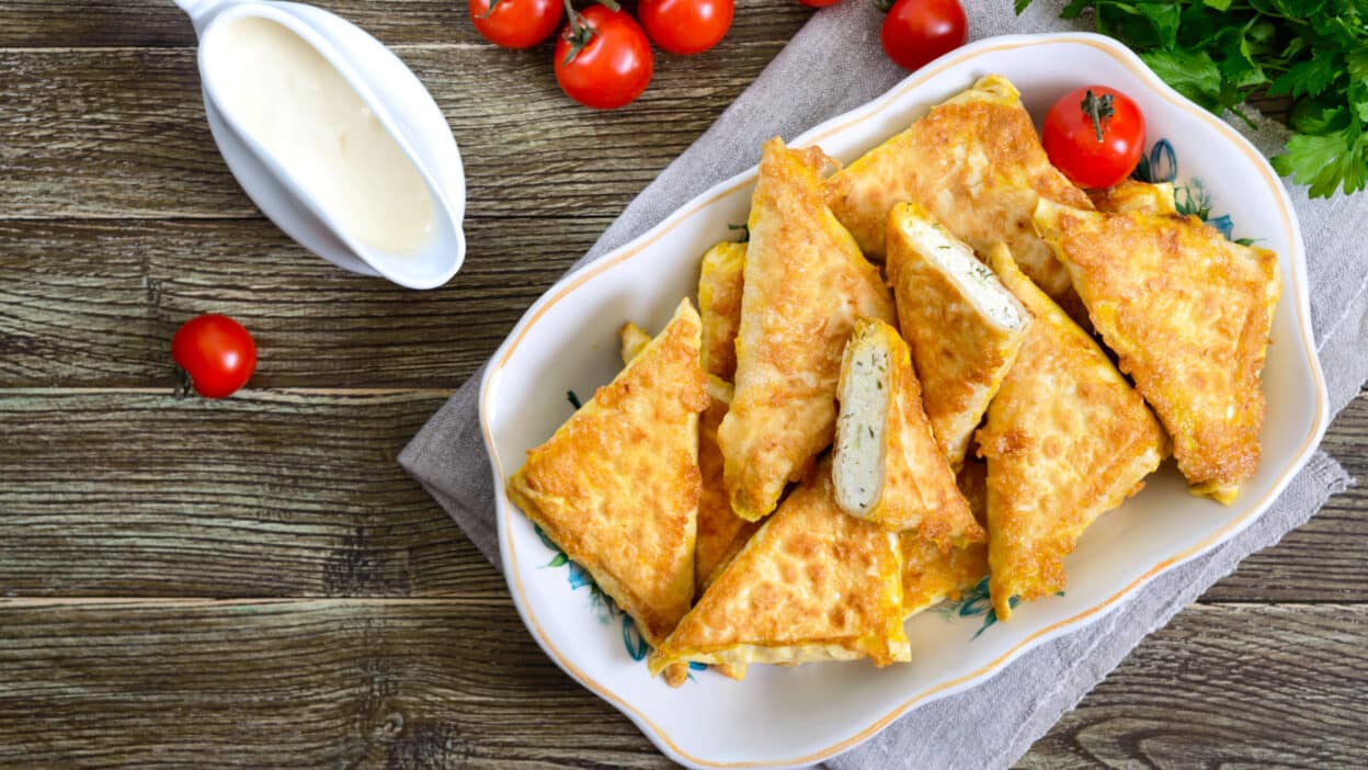 Golden fried zucchini triangles with creamy sauce, fresh cherry tomatoes, and parsley on a rustic wooden table.