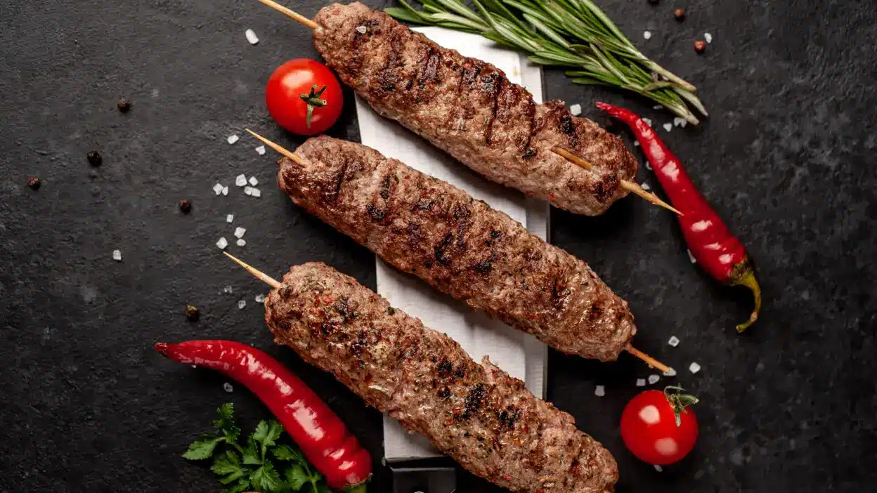 Grilled lamb kebabs on skewers with rosemary, cherry tomatoes, and red chili peppers on a dark surface.