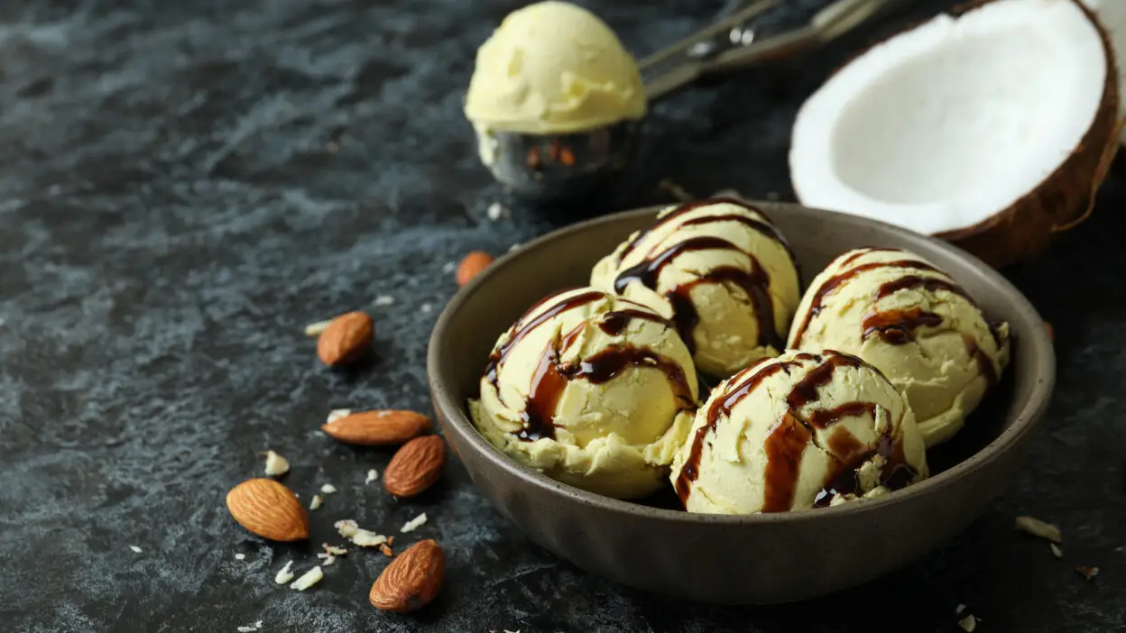 Coconut almond ice cream with chocolate drizzle in a bowl, surrounded by almonds and coconut on a dark surface.