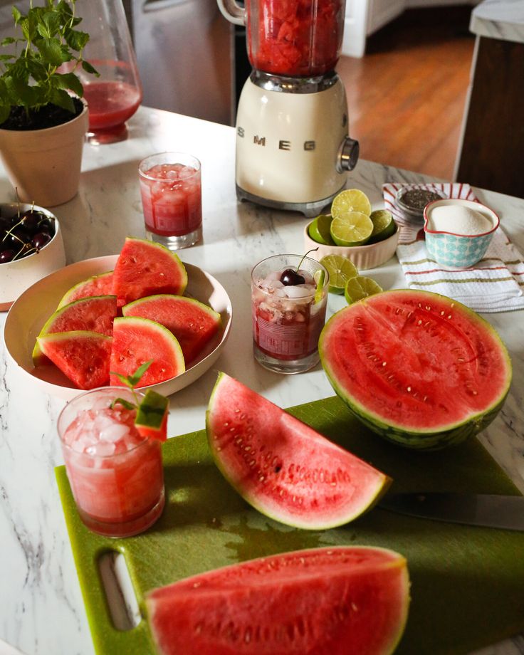 Fresh watermelon slices and smoothies on a kitchen counter with blender, limes, and cherries. Refreshing summer setup.