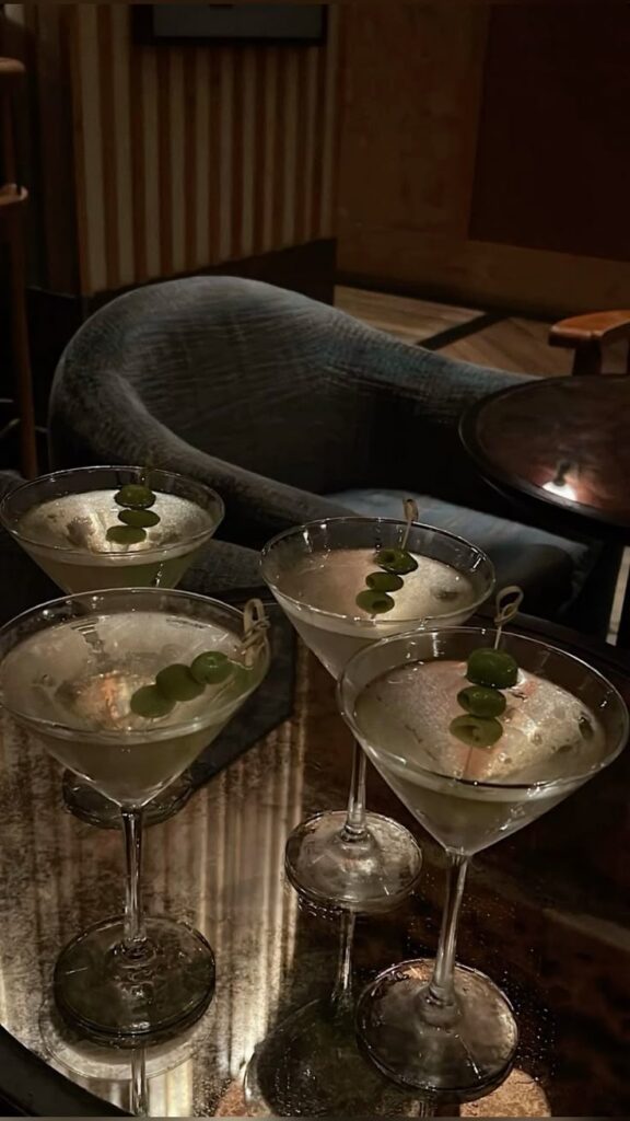 Four martinis with olives on a table in a cozy bar setting.