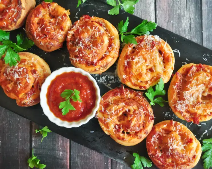 Pizza pinwheels with marinara sauce and parsley garnish on a black slate board, perfect for appetizers.