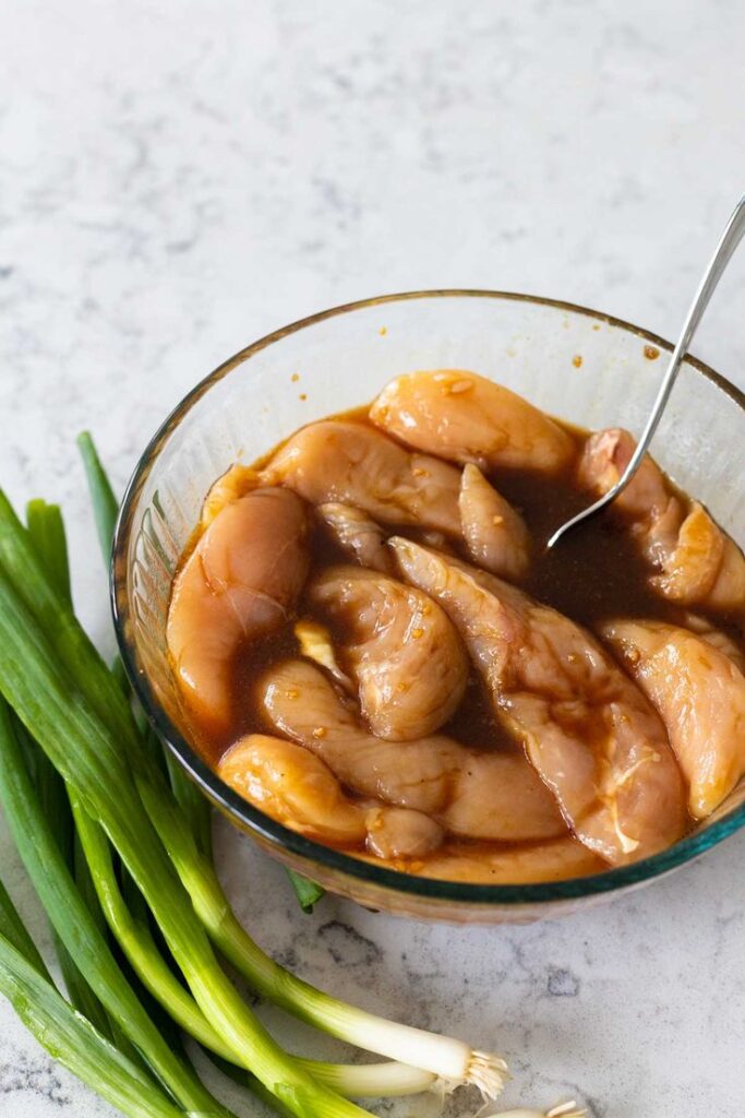 Marinated chicken breast in glass bowl with fresh green onions on marble surface.