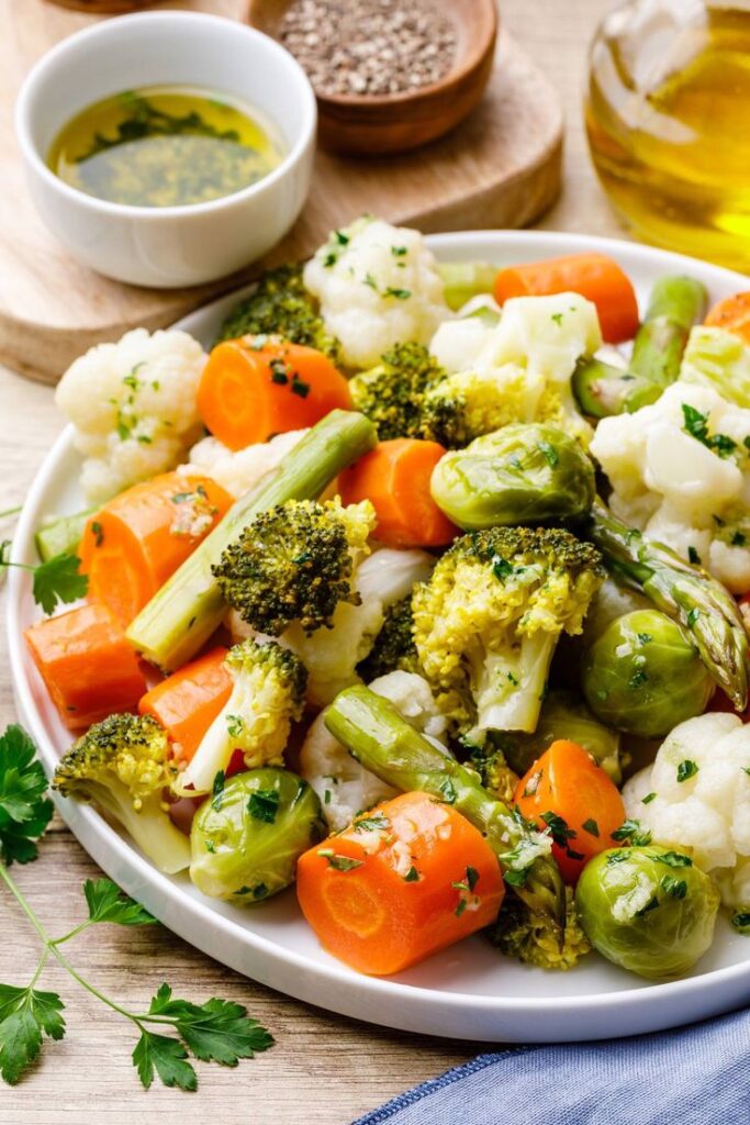 Assorted steamed vegetables including carrots, broccoli, and Brussels sprouts on a plate with herbs and olive oil.