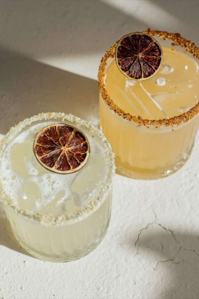 Two refreshing cocktails with dried lime garnishes and salt rims on a textured surface.