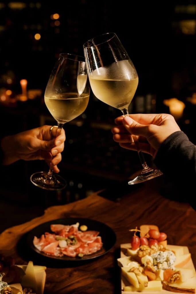 Two glasses of white wine clinking near a table with charcuterie and cheese, creating a cozy celebration atmosphere.