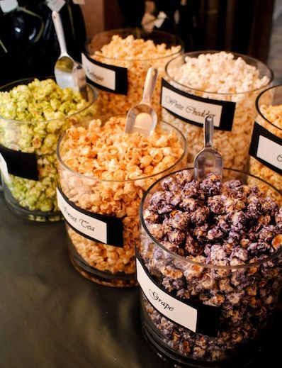 Assorted flavored popcorn in clear containers, including grape and green tea, displayed with scoops at a snack station.
