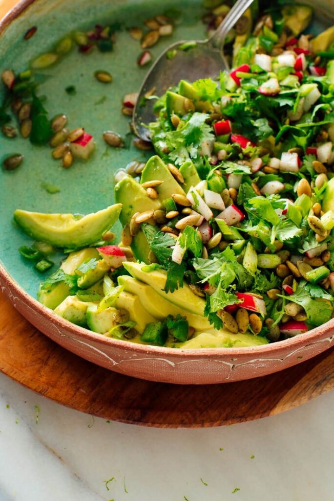 Fresh avocado salad with herbs, radish, and seeds in a ceramic bowl on a wooden board.