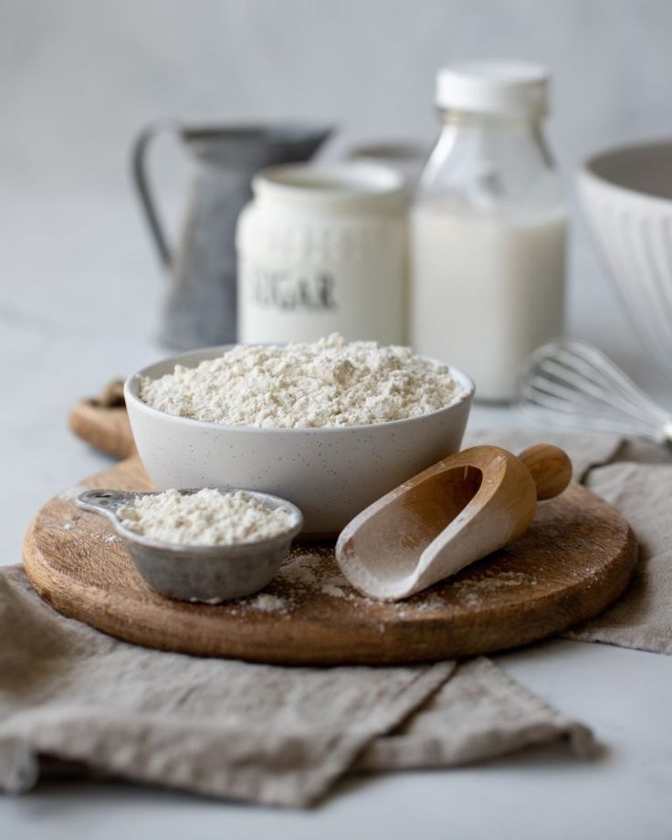 Bowl of flour with wooden scoop on rustic board, surrounded by baking ingredients like milk and sugar on a kitchen counter.