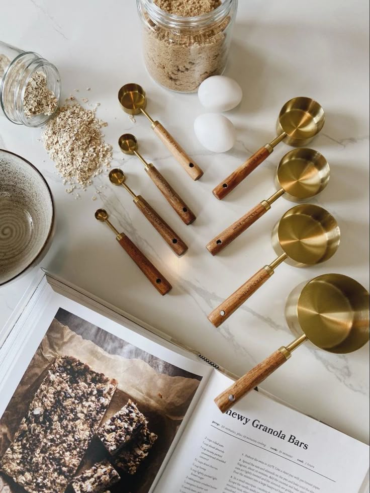 Granola bar recipe book with oats, brown sugar, eggs, and brass measuring spoons on a kitchen counter.