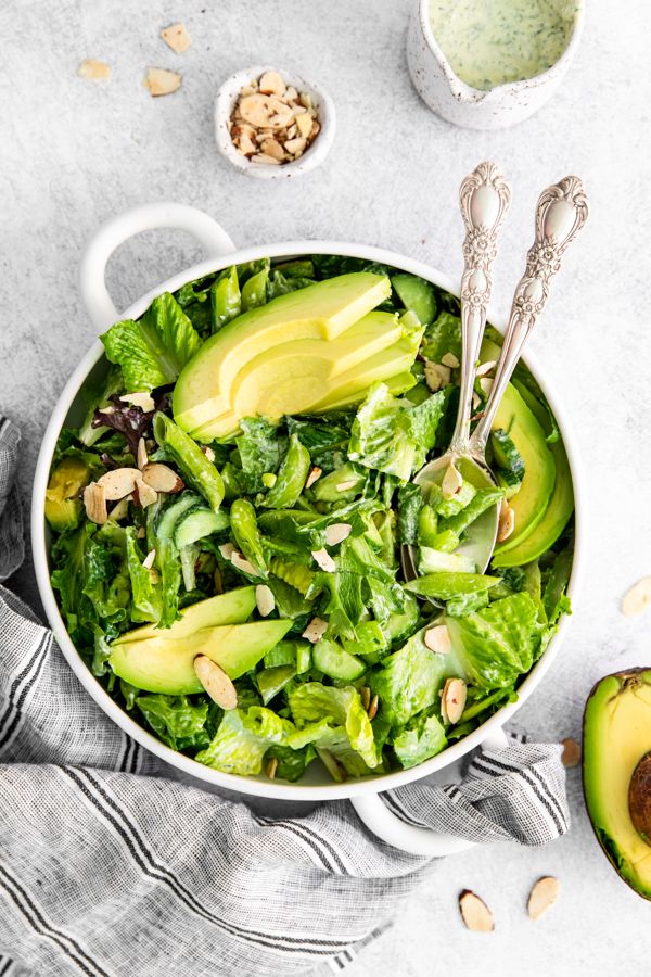 Fresh green salad with avocado, mixed greens, and sliced almonds in a white bowl, on a gray cloth napkin.