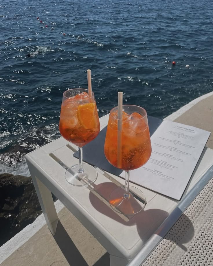 Two vibrant cocktails on a seaside table with a menu under the sun, overlooking a sparkling ocean.