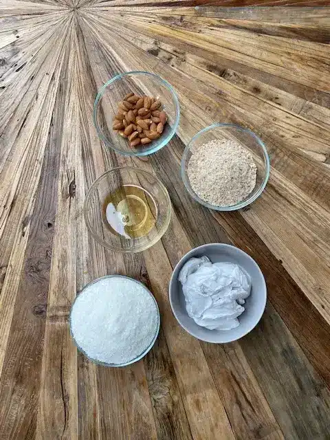 Ingredients for a homemade recipe: almonds, oats, sugar, coconut, oil, displayed on a wooden table surface.
