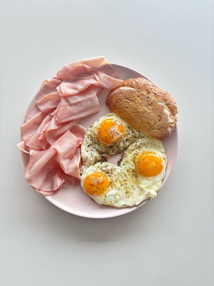 Breakfast plate with fried eggs, sliced ham, and a crusty bread roll on a pink dish.