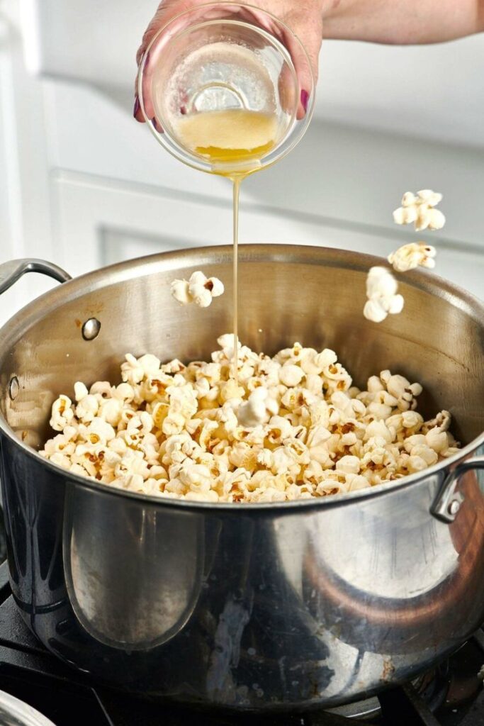 Pouring melted butter onto freshly popped popcorn in a stainless steel pot on the stove.