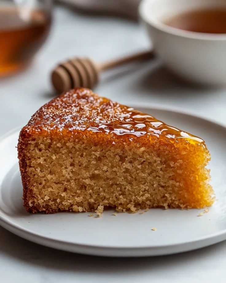 Slice of moist honey cake drizzled with syrup on a plate, accompanied by a honey dipper and tea cup in the background.