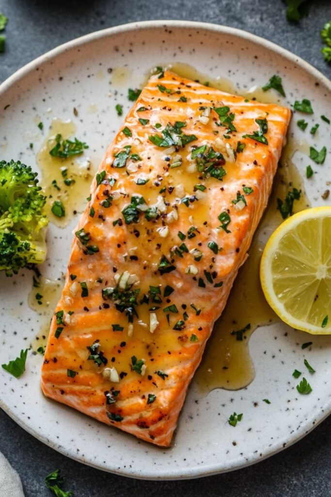 Grilled salmon fillet with herbs and lemon slice on a plate, garnished with broccoli and seasoning.