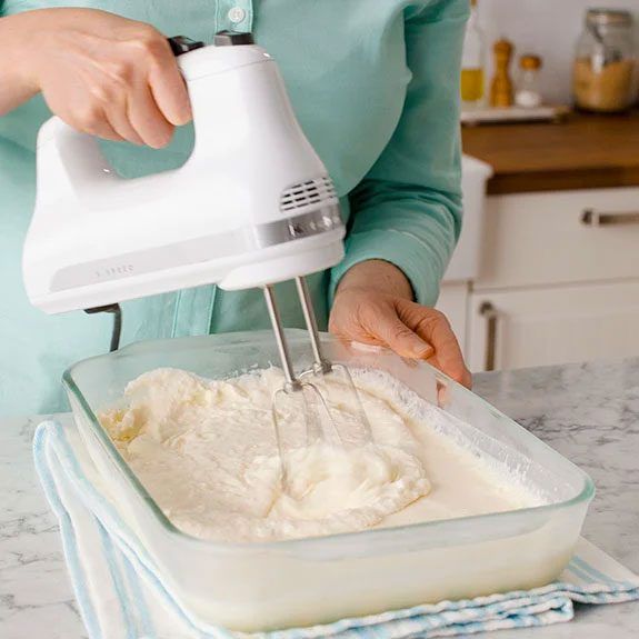 Person using a hand mixer to blend creamy batter in a glass dish on a kitchen counter.