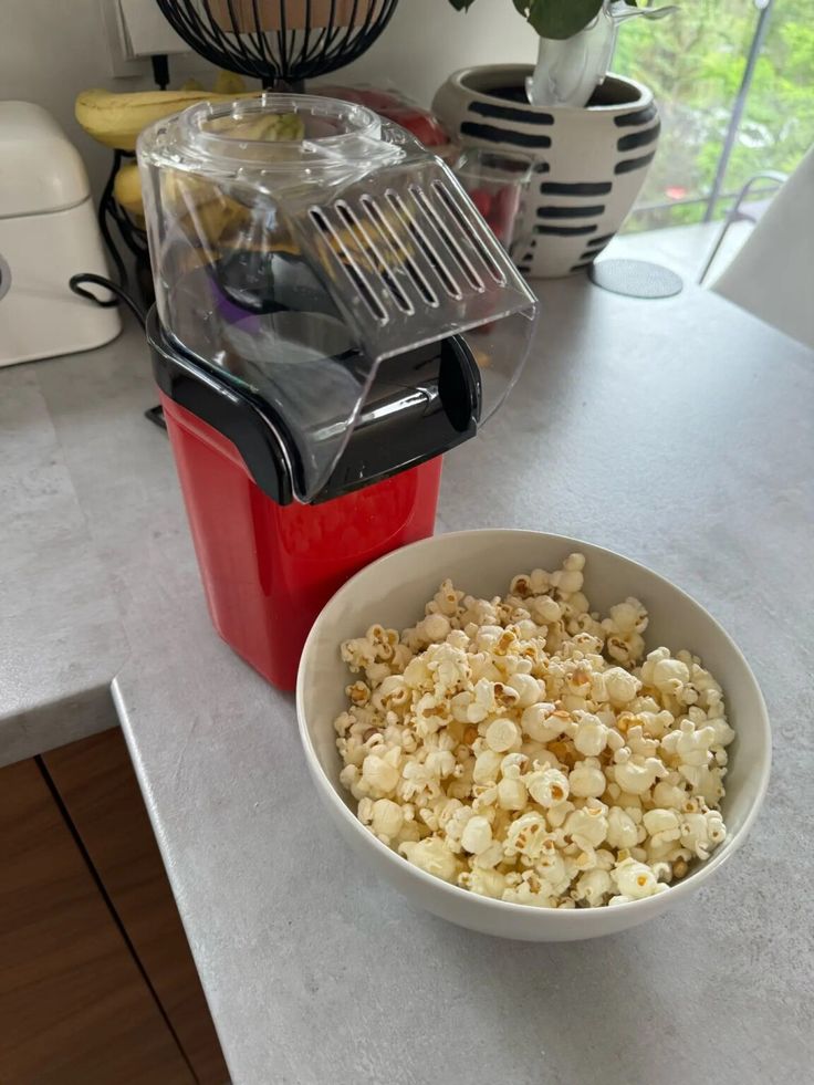 Bowl of fresh popcorn beside a red hot air popper on a kitchen counter.