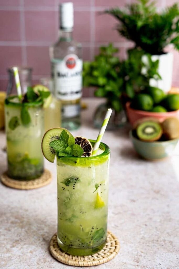 Refreshing kiwi mojito cocktail with mint leaves, limes, and a straw on a stone countertop with rum and fruit decor.