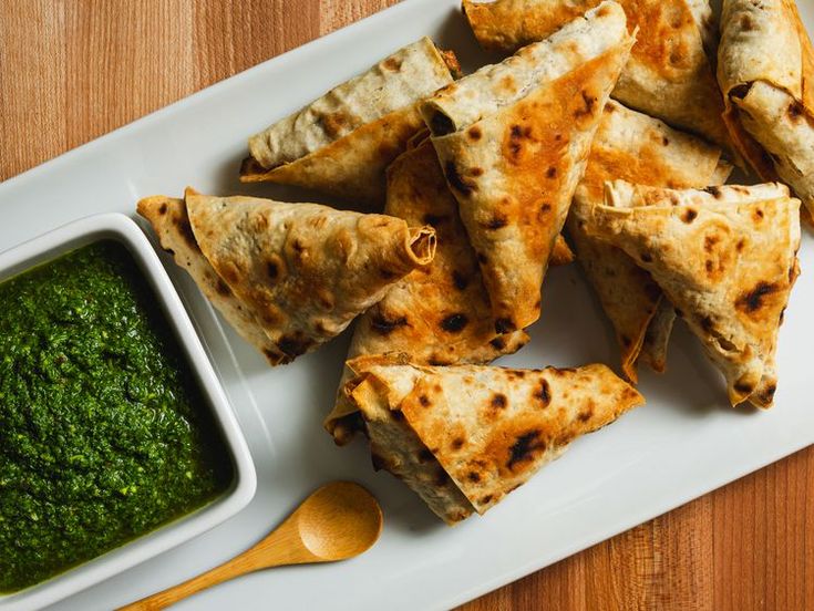 Crispy samosas with green chutney on a white platter, accompanied by a wooden spoon, on a wooden table.