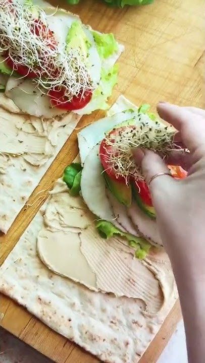 Making healthy wraps with hummus, turkey, lettuce, tomato, and sprouts on flatbread on a wooden cutting board.