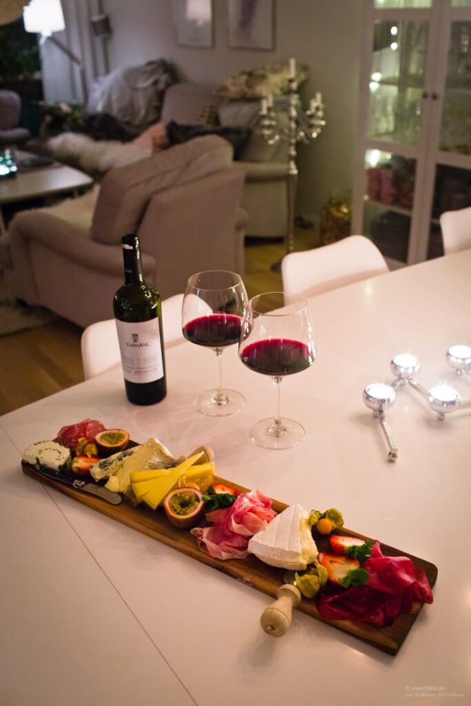 Charcuterie board with cheese, meats, fruit, and two glasses of red wine on a cozy dining table.