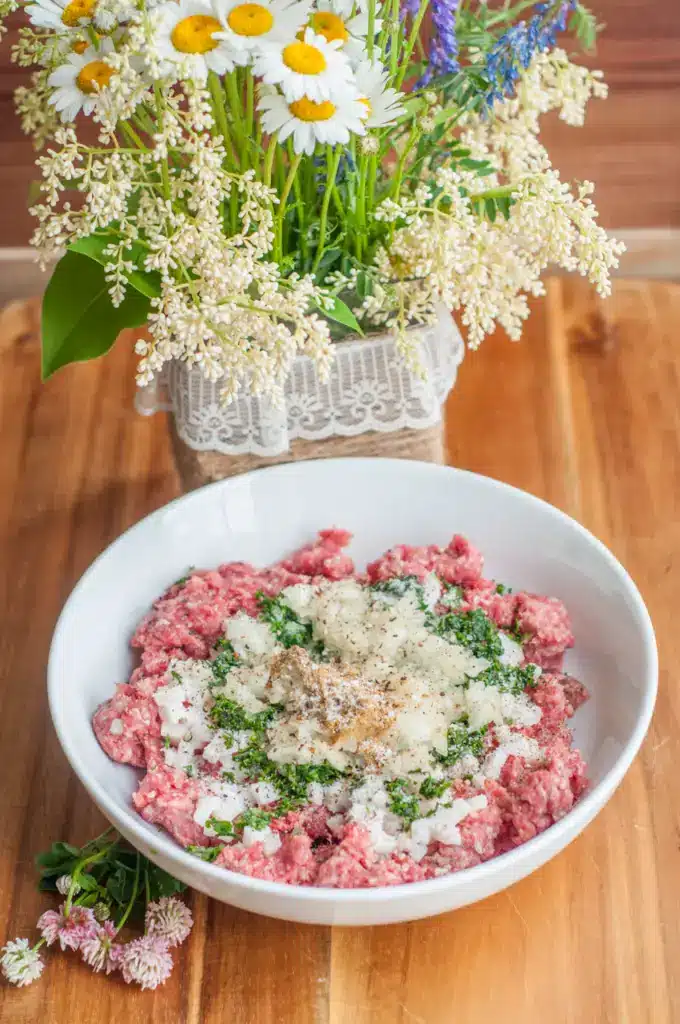 Minced meat with spices and herbs in a bowl, wooden table, floral centerpiece in the background.