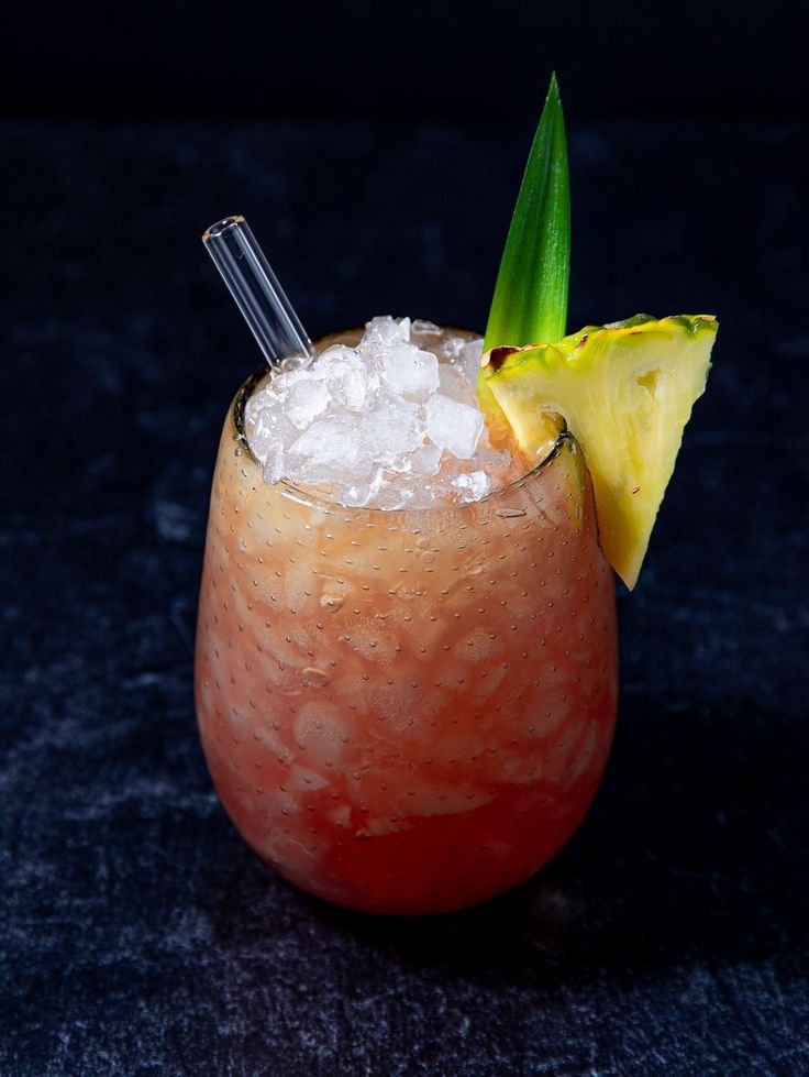 Tropical cocktail with ice, pineapple slice, and leaf garnish in a glass, set on a dark surface.