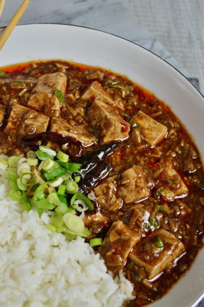 Spicy mapo tofu with sliced green onions and steamed rice on a white plate, garnished with dried red chilies.