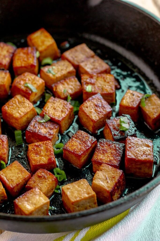 Golden-brown tofu cubes sizzling in a cast iron skillet with garnish. Vegan dish cooking close-up.