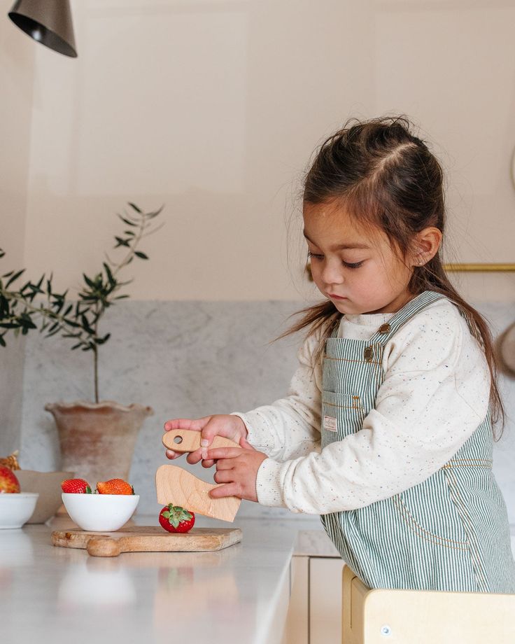 Child in overalls slicing strawberries in a kitchen with white bowls and a wooden board.