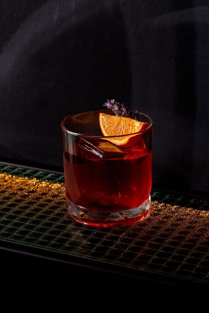 Red cocktail in a glass with an orange slice and lavender garnish on a reflective surface.