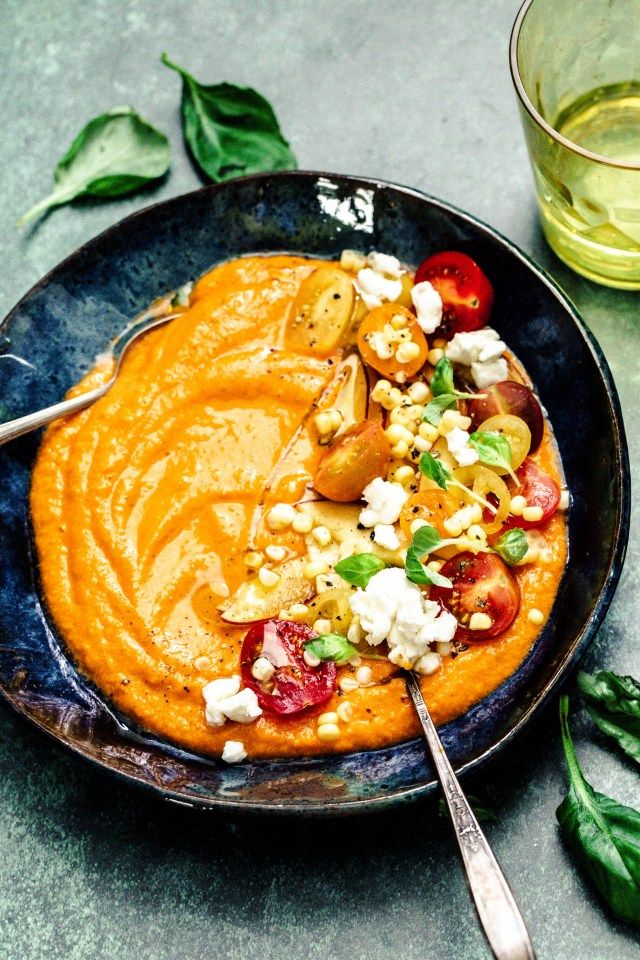 Creamy tomato soup in a bowl with cherry tomatoes, feta cheese, and herbs, served on a textured table.