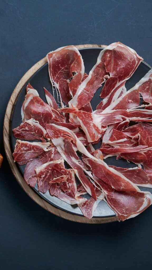 Plate of thinly sliced cured ham on a dark background, perfect for gourmet food blogs or charcuterie boards.
