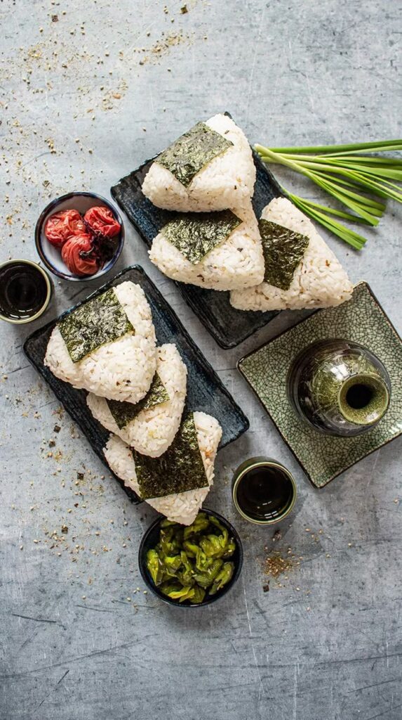 Onigiri rice balls with seaweed on plates, accompanied by pickles and beverages on a textured gray surface.