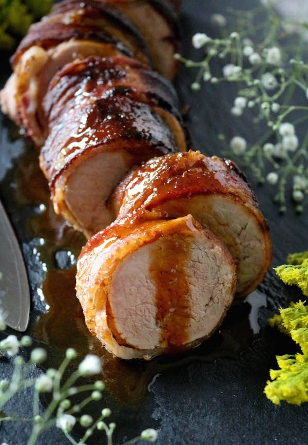 Sliced bacon-wrapped pork tenderloin on a black platter, garnished with fresh herbs, close-up.