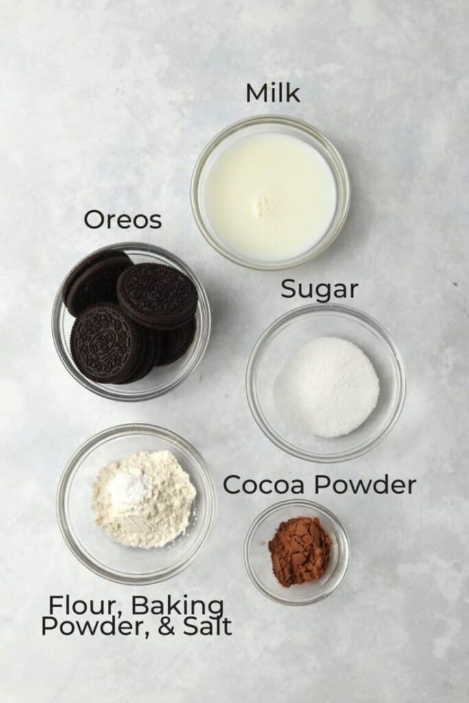 Ingredients for baking: oreos, milk, sugar, cocoa powder, flour, baking powder, and salt in bowls on a gray surface.