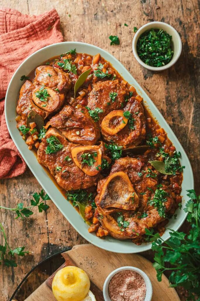 Plate of savory osso buco garnished with parsley on a rustic wooden table, accompanied by fresh herbs and lemon.