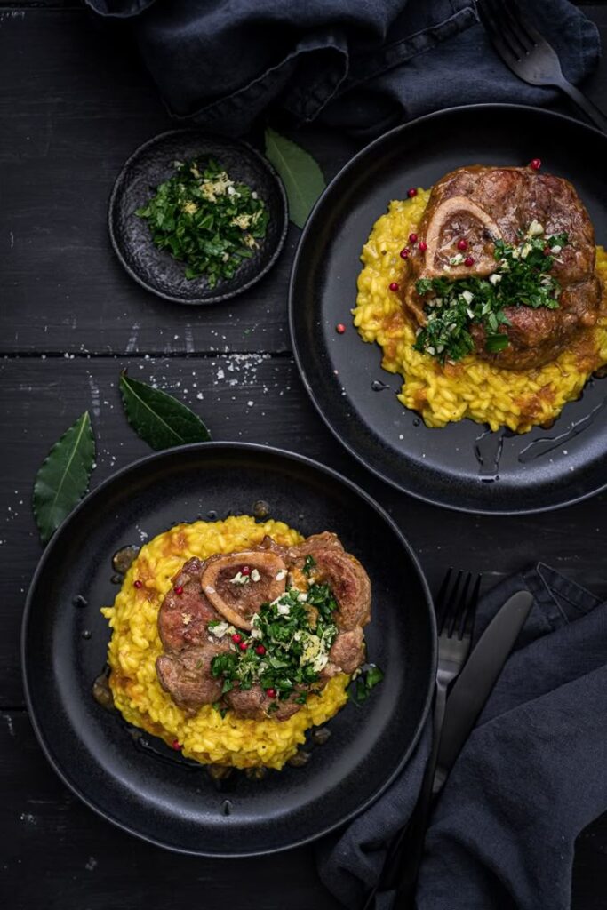 Two plates of osso buco with saffron risotto and gremolata garnish on a dark table setting.