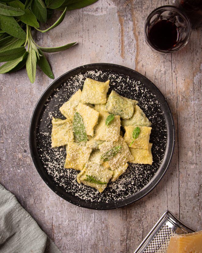 Plate of homemade ravioli with sage and grated cheese on a rustic table, paired with a glass of red wine.